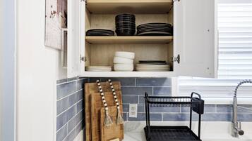 Una cocina con un armario que contiene platos y tazones, una tabla de cortar de madera y un escurreplatos negro.