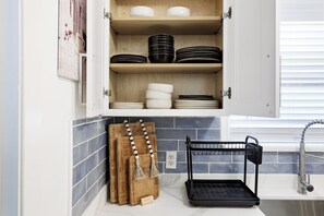 Una cocina con un armario que contiene platos y tazones, una tabla de cortar de madera y un escurreplatos negro.