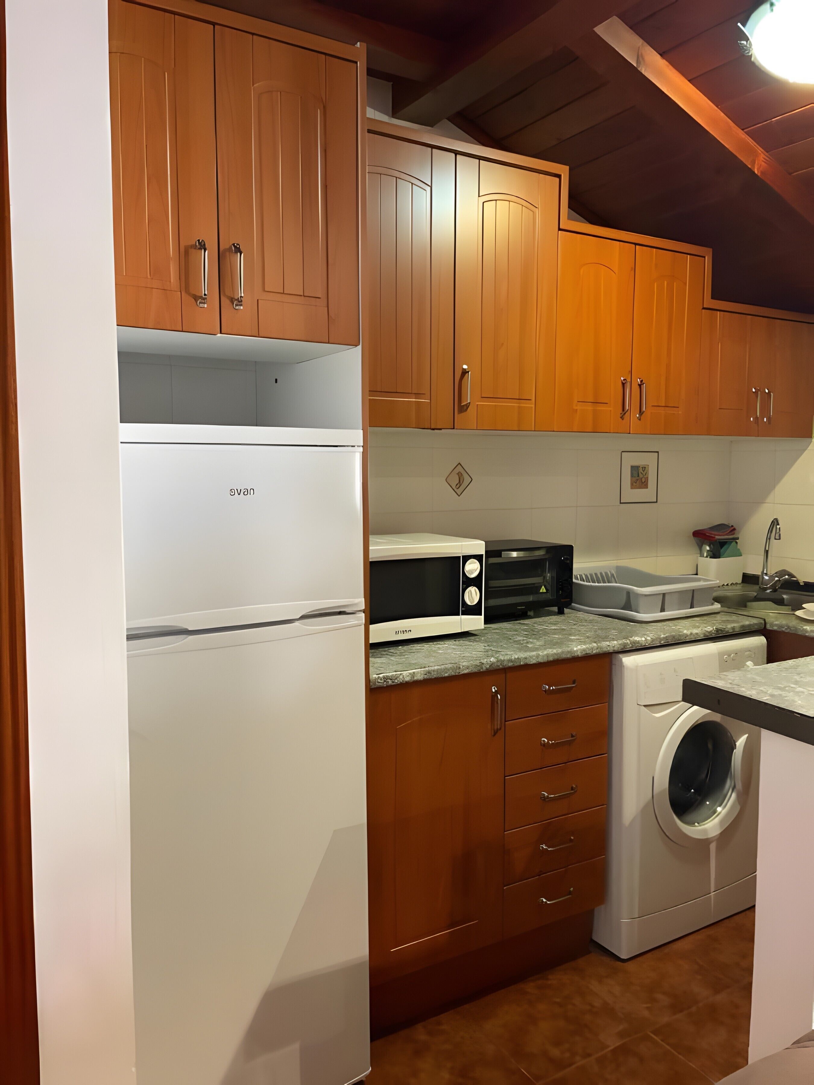 Een keuken met houten kasten, een witte koelkast, een magnetron, een vaatwasser en een wasmachine.