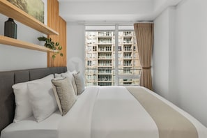A modern bedroom with a large bed, a wooden shelf with a plant, and a view of high-rise buildings. - Casanoma by Kozystay - Paskal (Kota Bandung)