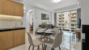A modern kitchen with wooden cabinets, a dining area with a round table and chairs, and a view of a residential building through large windows. - Casanoma by Kozystay - Paskal (Kota Bandung)