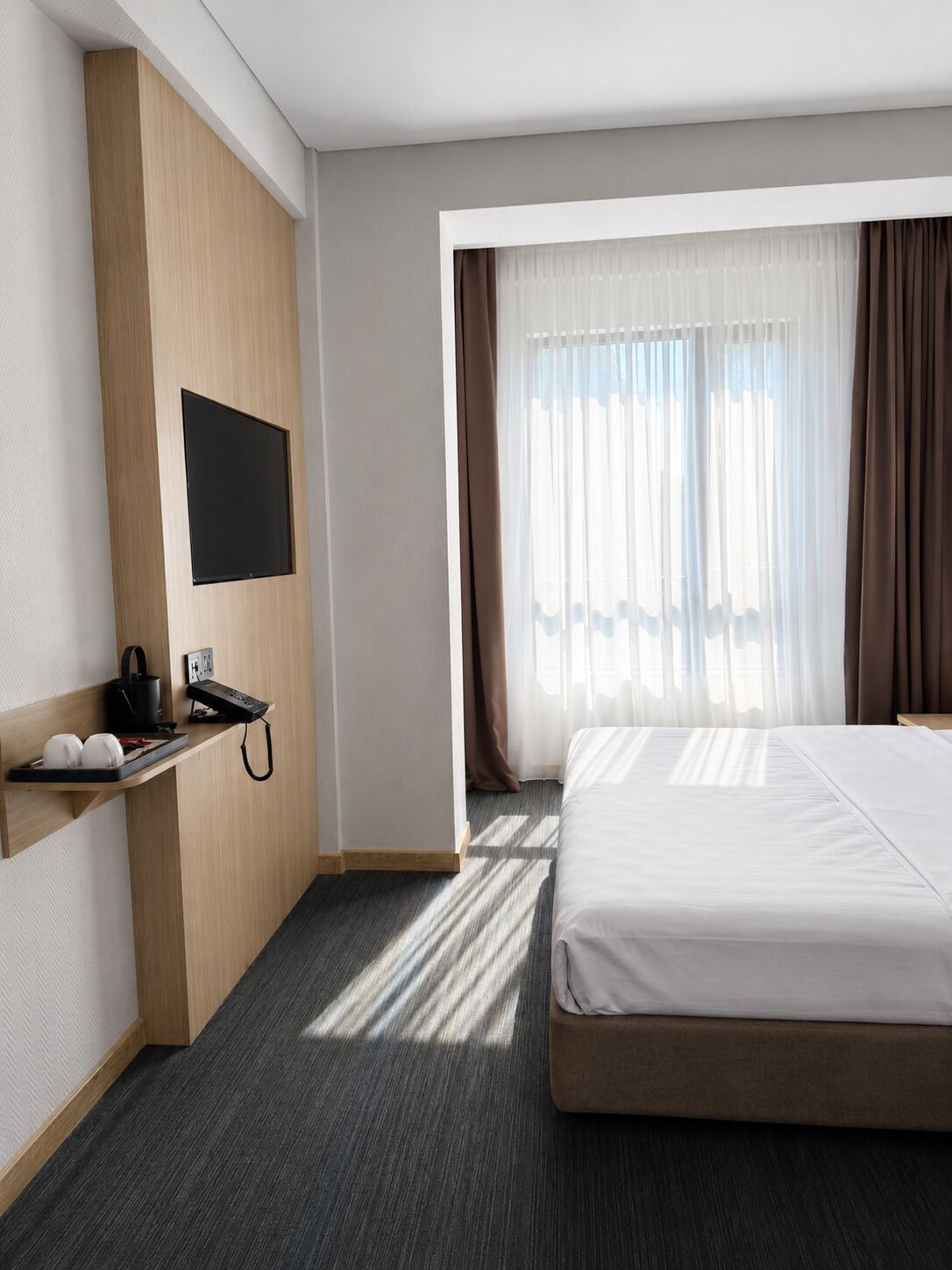 Habitación de hotel con cama, escritorio con teléfono, televisor de pantalla plana en la pared y ventana con cortinas.