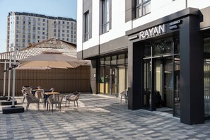 Exterior de un hotel con mesas al aire libre, fachada moderna y vistas a la ciudad.