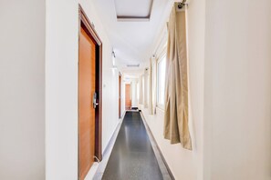 Corredor de hotel com uma porta de madeira à esquerda, cortinas bege à direita e piso de azulejos.