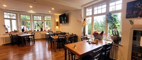 A dining area with wooden tables and chairs, large windows with views of greenery, and a wall-mounted flat-screen TV.