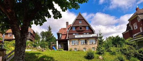 En traditionell byggnad med en skylt som visar texten ”HOTEL ROSENHOF”, belägen på landsbygden med en stenmur, grönska och en klar himmel.