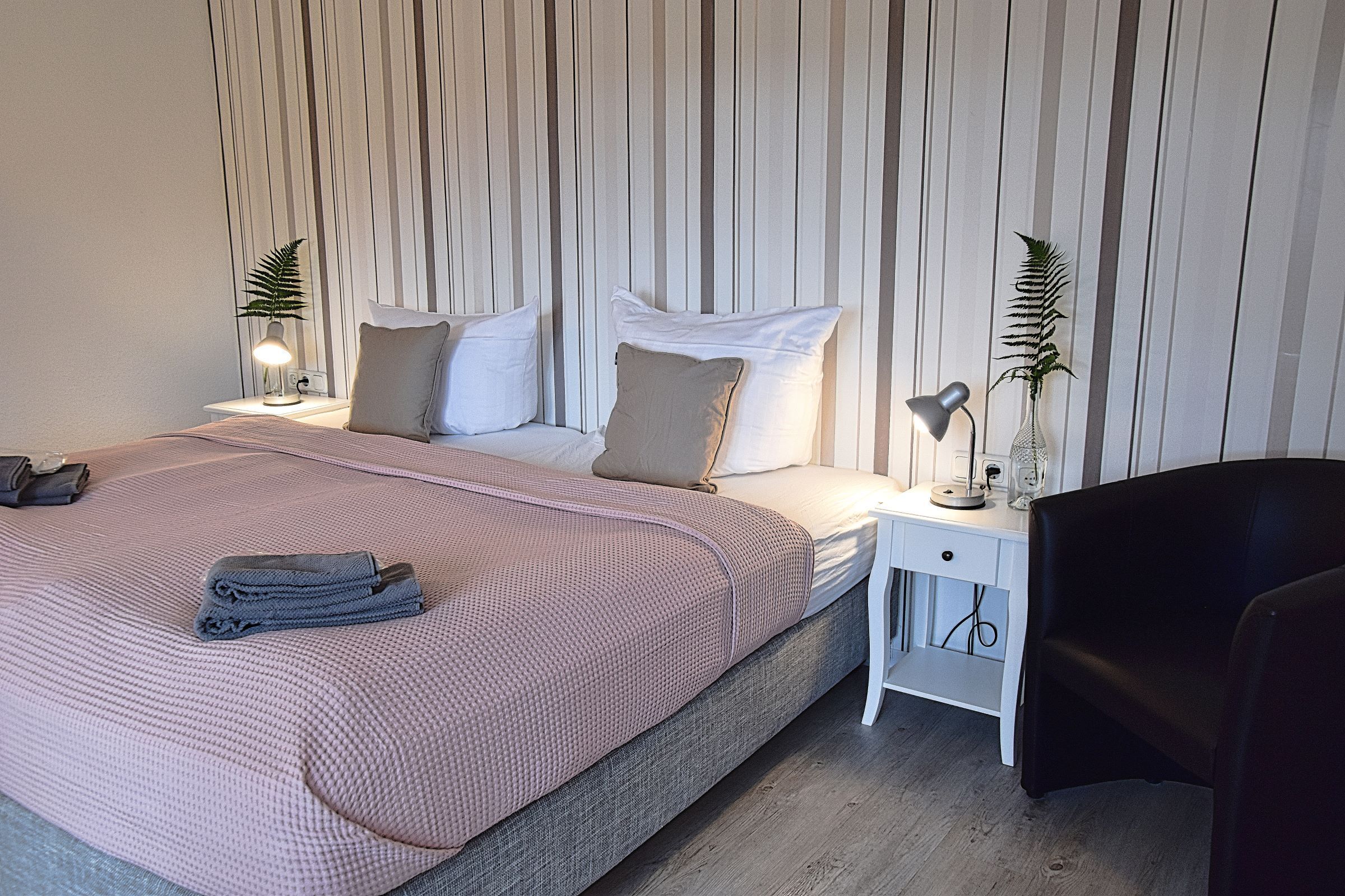 A neatly made bed with a pink blanket and pillows, a bedside table with a lamp, a chair, and a plant on the nightstand.