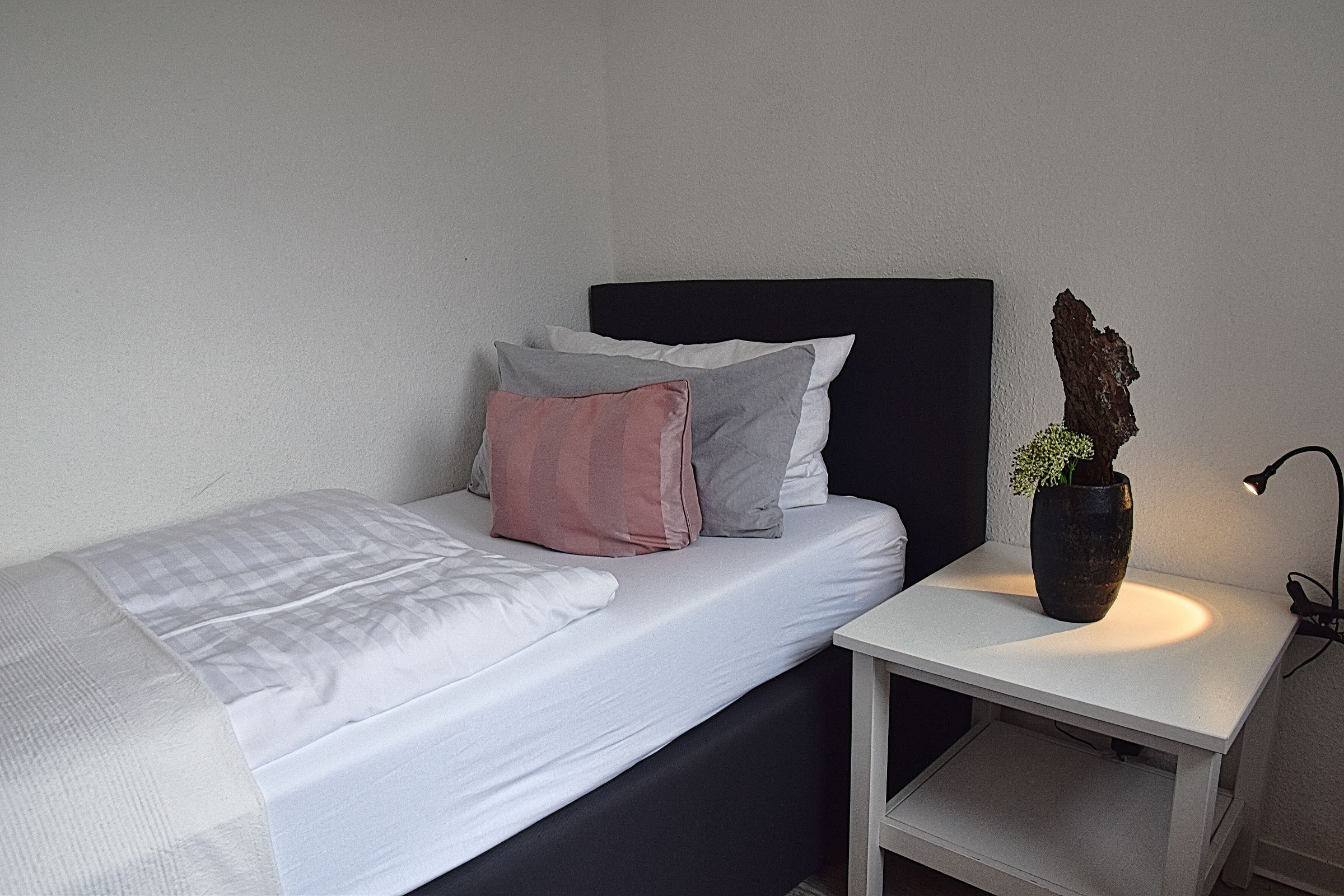 Een eenpersoonsbed met een witte matras en meerdere kussens in tinten grijs en roze. Een klein nachtkastje met een lamp en een vaas met een plantje staat naast het bed.