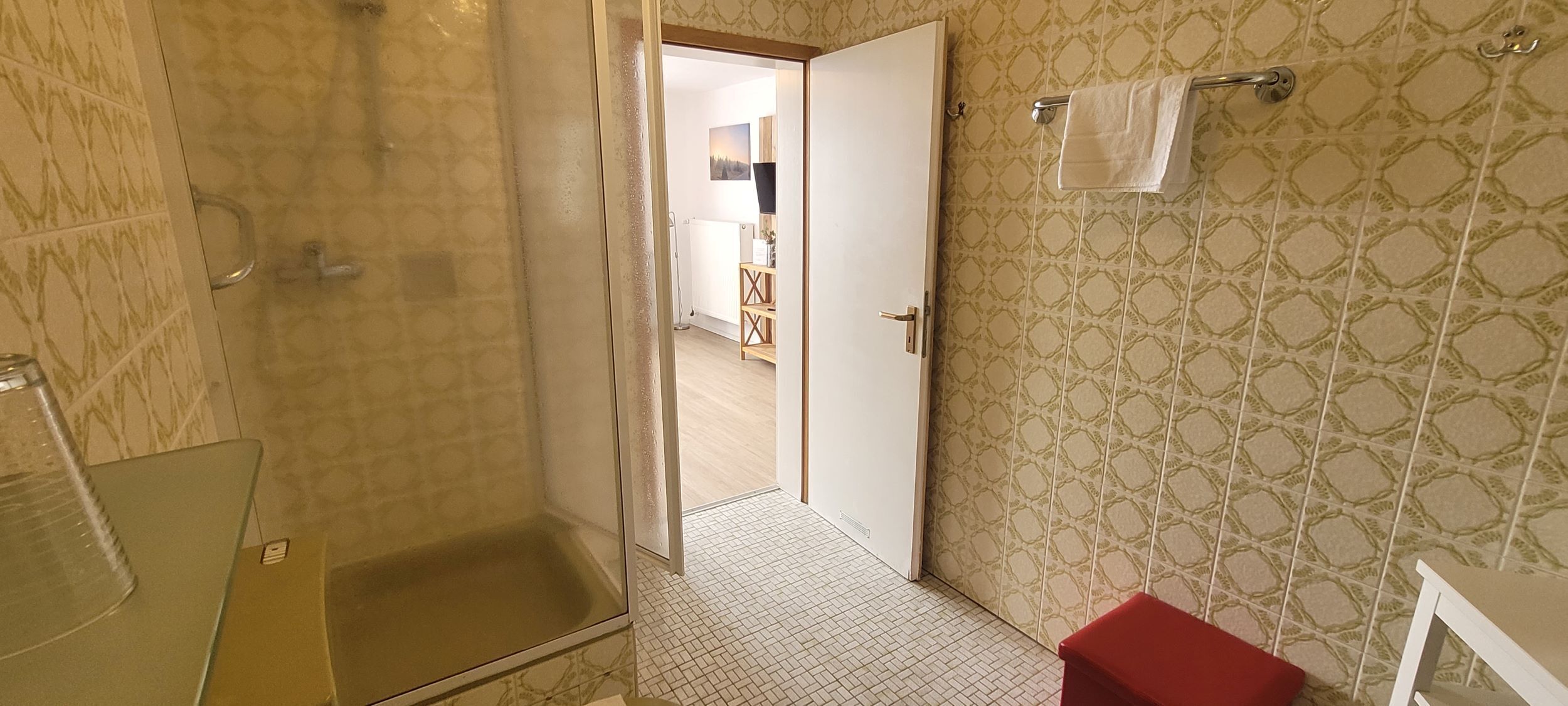 Baño con ducha, inodoro y lavabo. Hay una toalla colgada en un perchero y un taburete rojo.