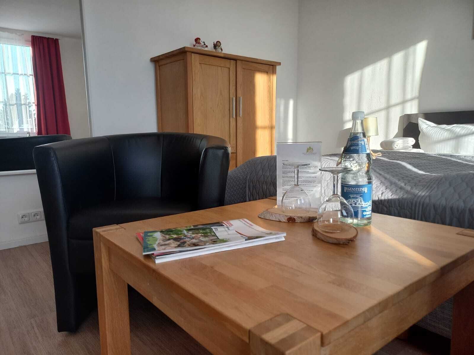 Ein Hotelzimmer mit Holztisch, schwarzem Ledersessel, Bett mit grauer Bettdecke, Holzschrank und Fenster mit roten Vorhängen.