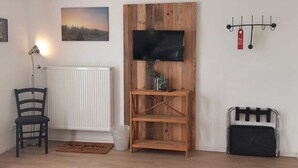 Una habitación con un radiador blanco, un mueble bajo de madera para televisor con una pantalla plana, una silla y un estante pequeño con una planta.