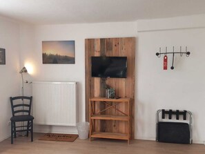 Una stanza con termosifone bianco, un mobiletto TV in legno con televisore piatto, una sedia e un piccolo ripiano con una pianta.
