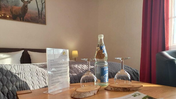 Habitación de hotel con cama, una mesa de madera con un menú, una botella de licor y una revista.