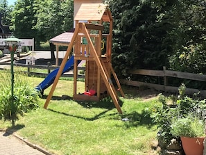 A wooden swing set with a slide in a grassy area, surrounded by a wooden fence and potted plants. - Double Room with Balcony (Braunlage)
