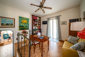A living room with a wooden floor, a wooden dining table, a yellow sofa, a bookshelf, and a ceiling fan. - Casa Serena: with Pine Forest, Jacuzzi Tub & BBQ near the beach, Pet Friendly (San Vito dei Normanni)
