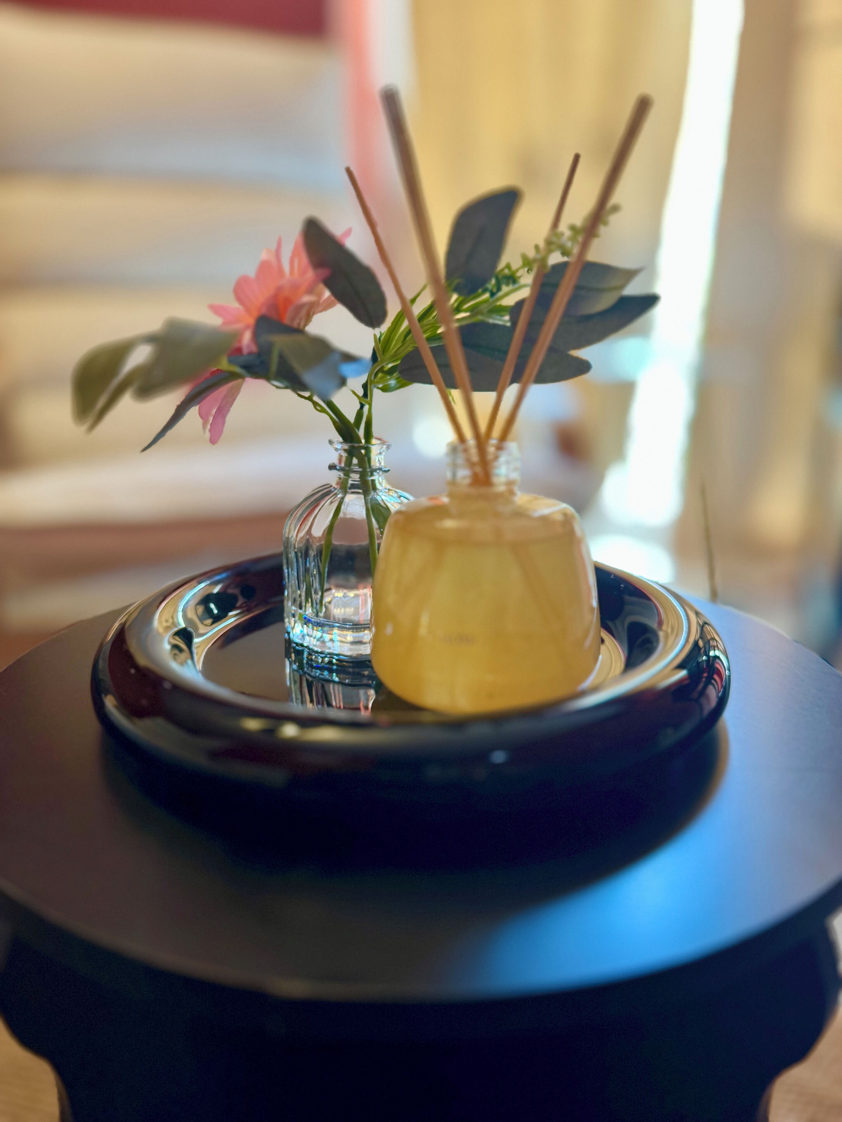 Ein kleiner Tisch mit einer Glasvase, die eine rosa Blume enthält, und einer Glasflasche mit Schilfrohlingen.