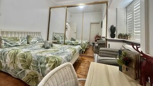 Hotellihuone, jossa on suuri sänky, oleskelualue pöydällä ja tuoleilla, peili ja ikkuna, jossa on sälekaihtimet.