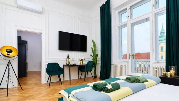 A hotel room with a bed, a desk, a chair, a television, and a view of a building outside. - Luxury Riverside Hideaway w/ Panoramic Views & Top Amenities – 622 (Budapest)