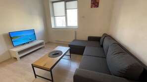 A modern living room with a flat-screen TV, a wooden coffee table, a grey sectional sofa, and a window with blinds.