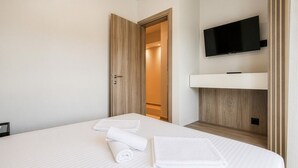 A hotel room with a bed, a TV mounted on the wall, and a wooden door leading to a hallway. - Sani Astera Deluxe by Rentals Pro - Sani Halkidiki (Sani)
