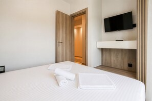 A hotel room with a bed, a TV mounted on the wall, and a wooden door leading to a hallway. - Sani Astera Deluxe by Rentals Pro - Sani Halkidiki (Sani)