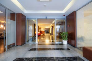 A hotel lobby with a glass entrance, a reception desk, seating area, and a potted plant.