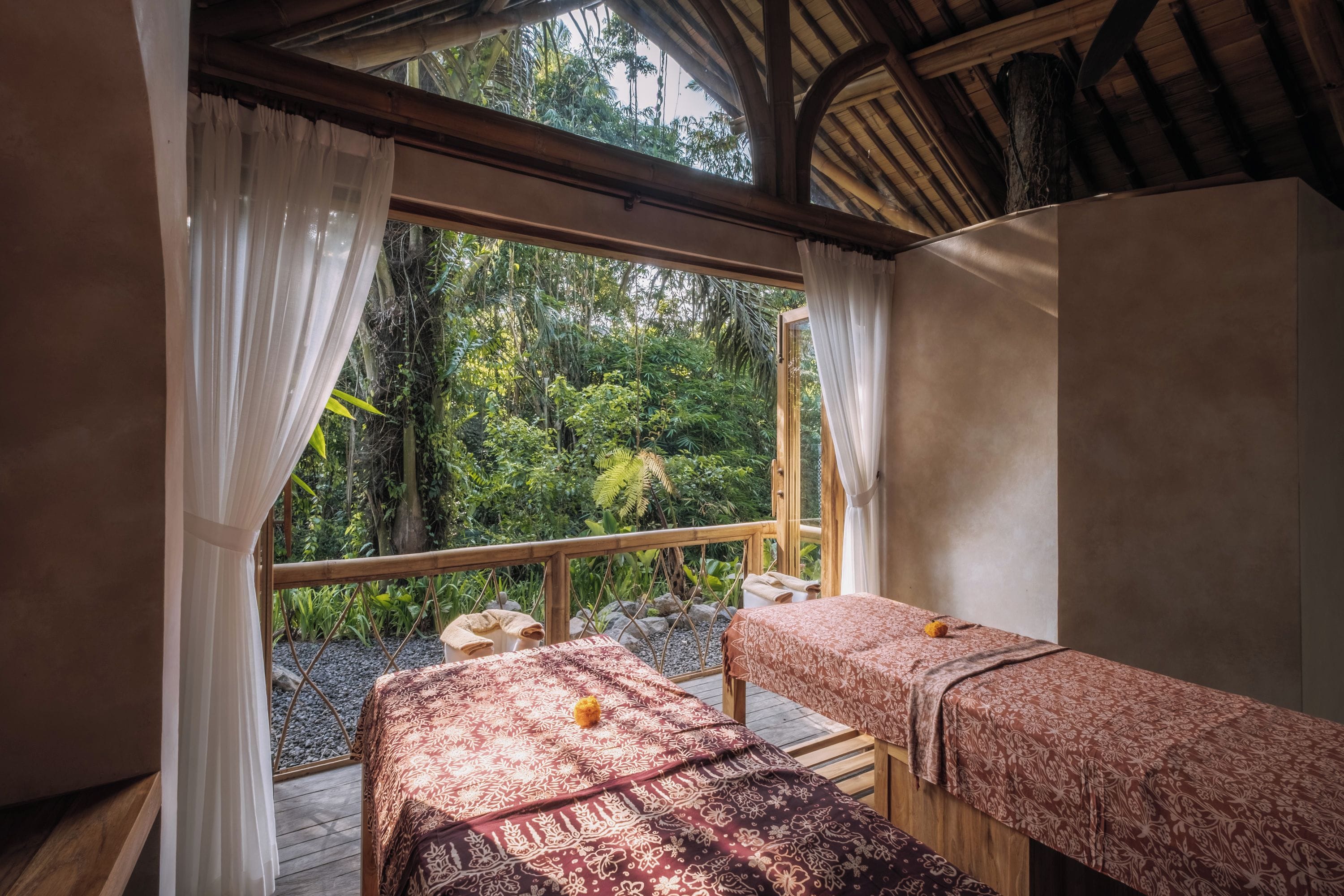 A room with two beds, a window with curtains, and a view of greenery outside.