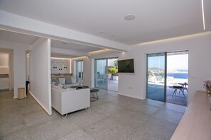 A modern interior with a white sofa, a dining area, and a view through large glass doors to an outdoor terrace. - Mykonos Seabreeze Town Villa (Mykonos)