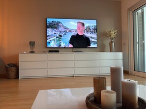 Ein Wohnzimmer mit einem an der Wand befestigten Flachbildfernseher, einer weißen Unterhaltungsanlage darunter und einem Fenster zur rechten Seite.