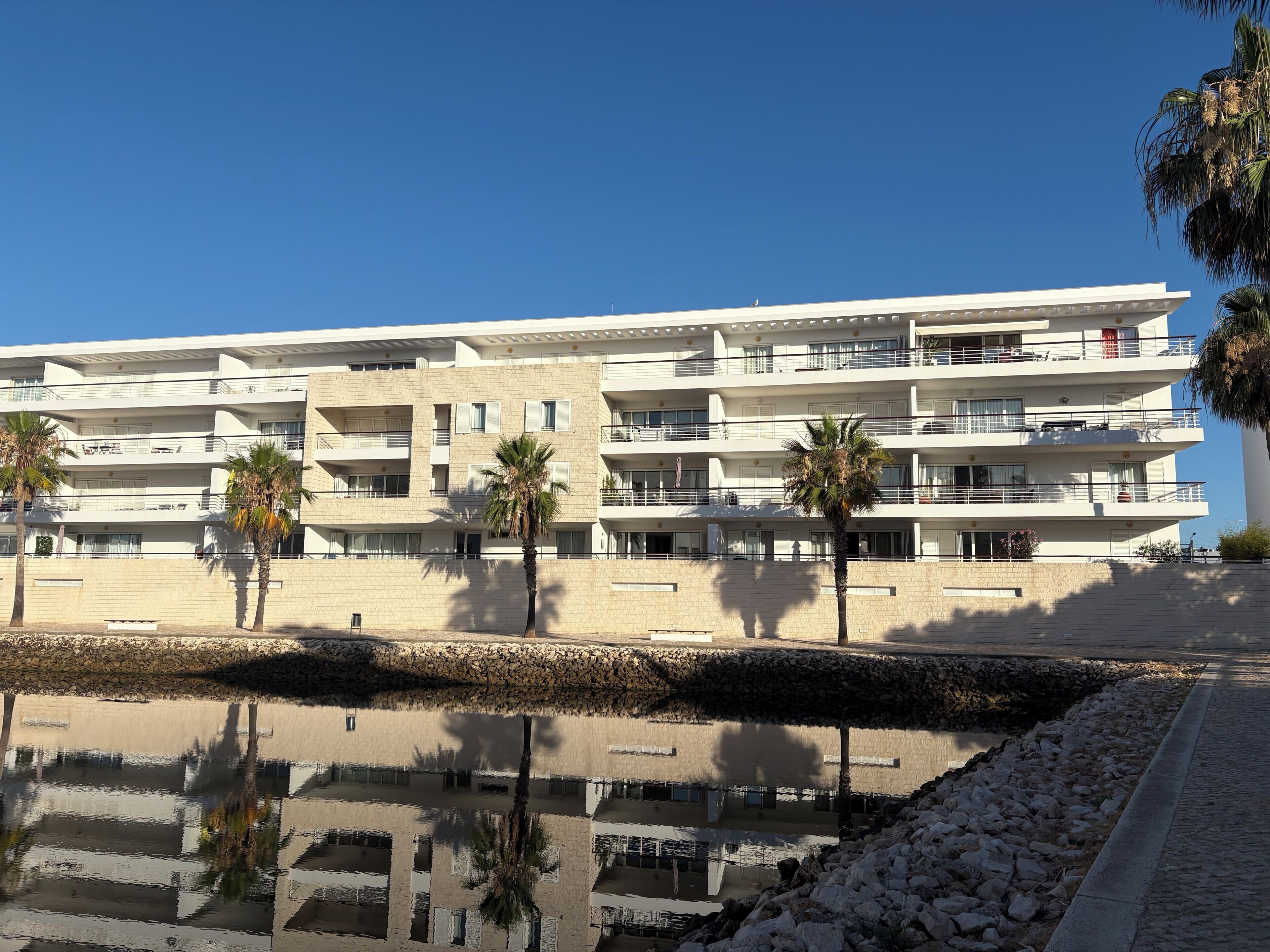 A multi-story hotel with balconies, palm trees, and a reflection in the water.