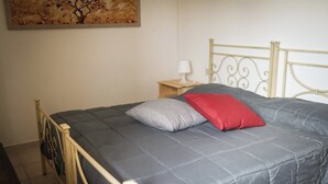 A bed with a metal frame, a grey quilt, and two pillows, one grey and one red.