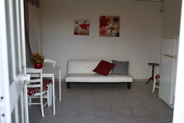 A small, clean room with a white sofa, a red pillow, a dining table with chairs, and two wall-mounted paintings.