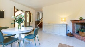 A dining area with a round table and chairs, a kitchen with white cabinets, a fireplace, and a staircase leading to another level. - Villa Grazia – Your relaxing escape just steps from the sea 🌿☀️ (Santa Margherita di Pula)