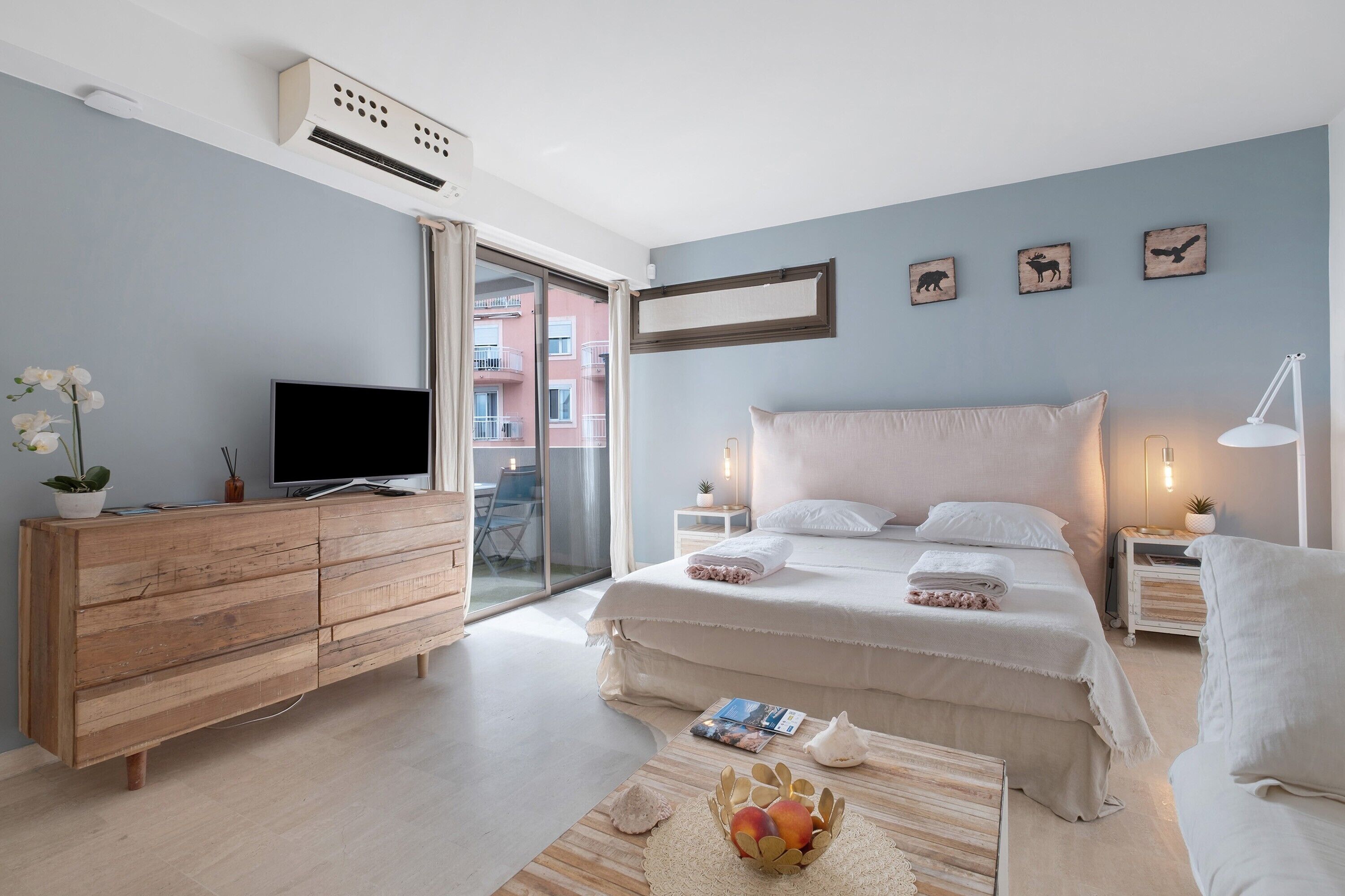 Ein modernes Schlafzimmer mit einem Bett, einem Fernseher, einer hölzernen Kommode und Blick auf ein Gebäude durch eine Glastür.