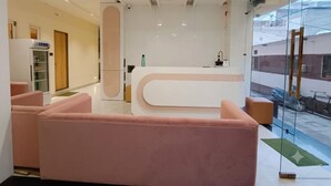 A modern reception area with a curved reception desk, a seating area with pink sofas, and a glass wall offering a view of the outside. - Hotel Agrawal Inn (Ujjain)