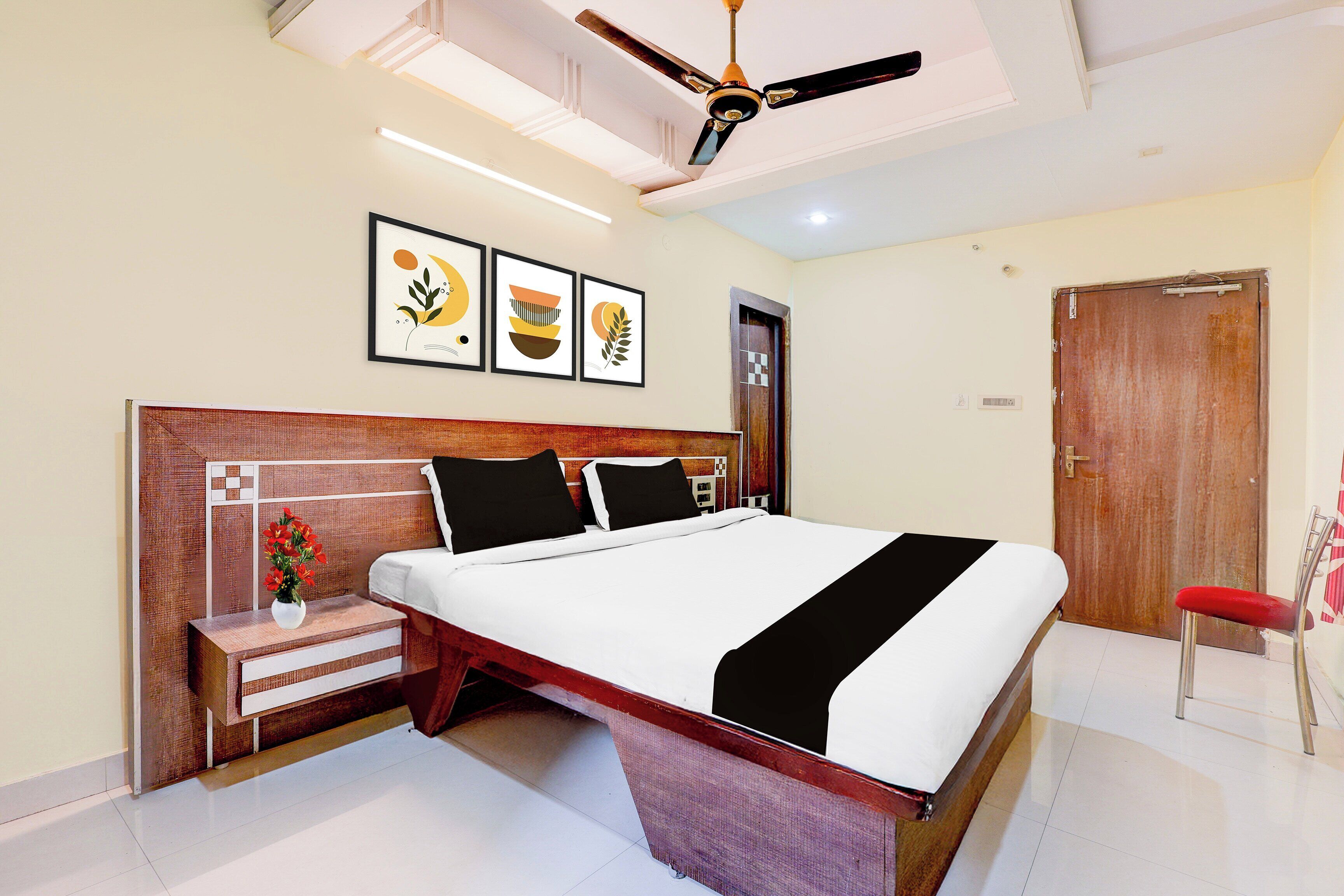 A hotel room with a bed, a nightstand with a vase, a ceiling fan, and framed artwork on the wall.