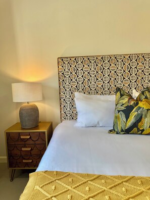 Un lit bien fait, avec une literie blanche, une tête de lit à motifs, une lampe de chevet et une table de chevet en bois dotée de tiroirs.
