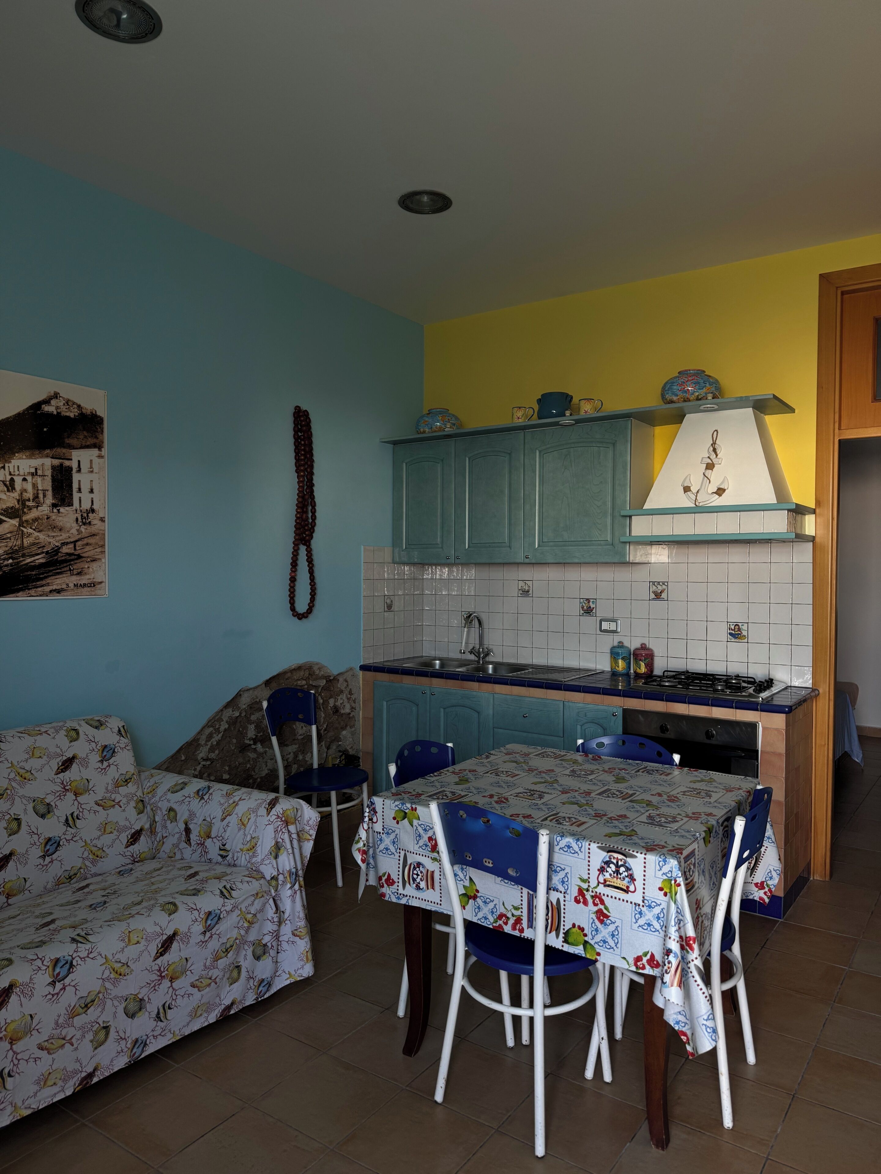 Een compacte keuken met blauwgroene kastjes, een betegelde spatwand en een eethoek met een tafel gedekt voor twee personen.