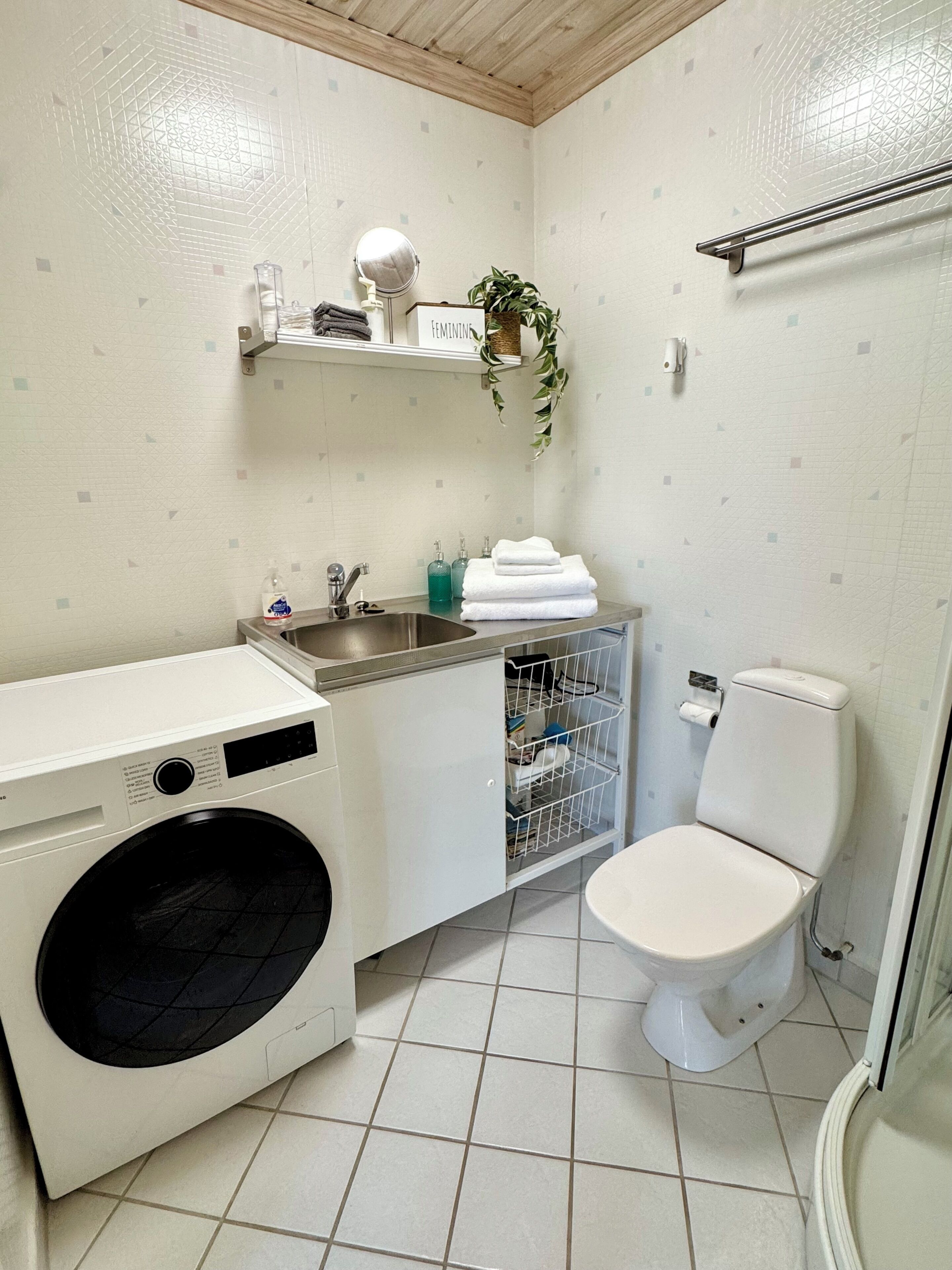 Ett litet badrum med tvättmaskin, toalett, handfat och en hylla med toalettartiklar.