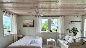 A bedroom with a bed, a sofa, a small table, and a ceiling fan. There are windows with curtains, a view of greenery outside, and a framed picture on the wall. - GG Village (Askim)