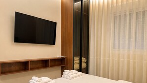 Et hotelværelse med en seng, et fladskærms-TV ophængt på væggen, en træhylde og et vindue med lette gardiner.