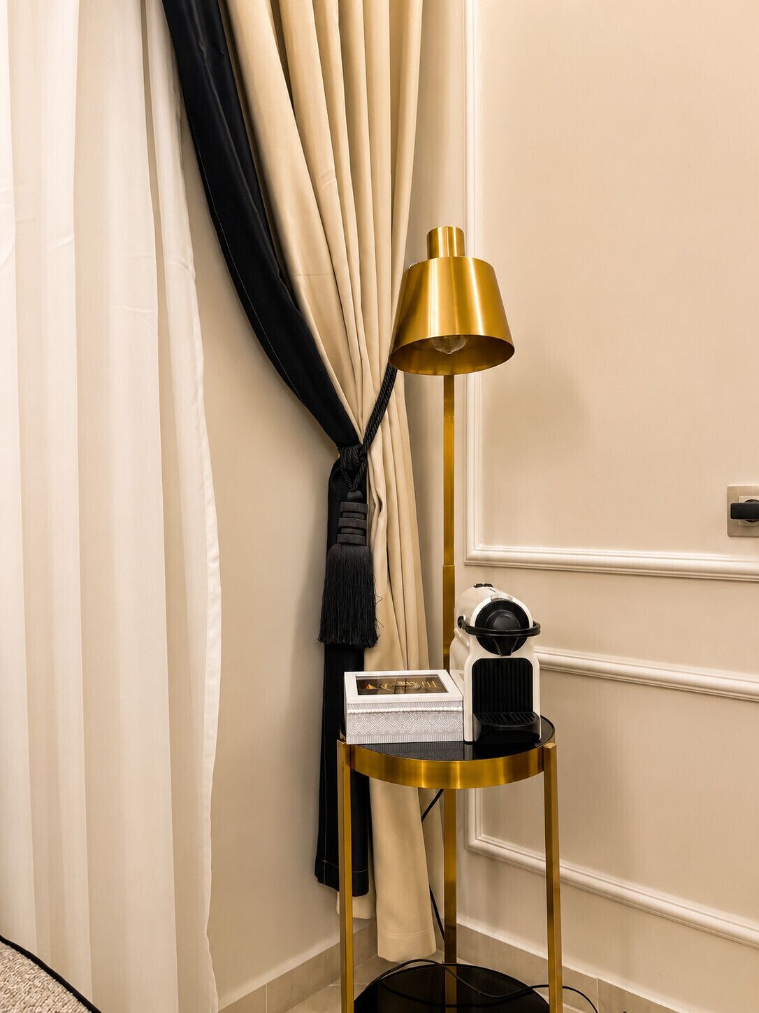 Rincón de una habitación con una lámpara de pie, una mesita con un objeto decorativo y una cortina.