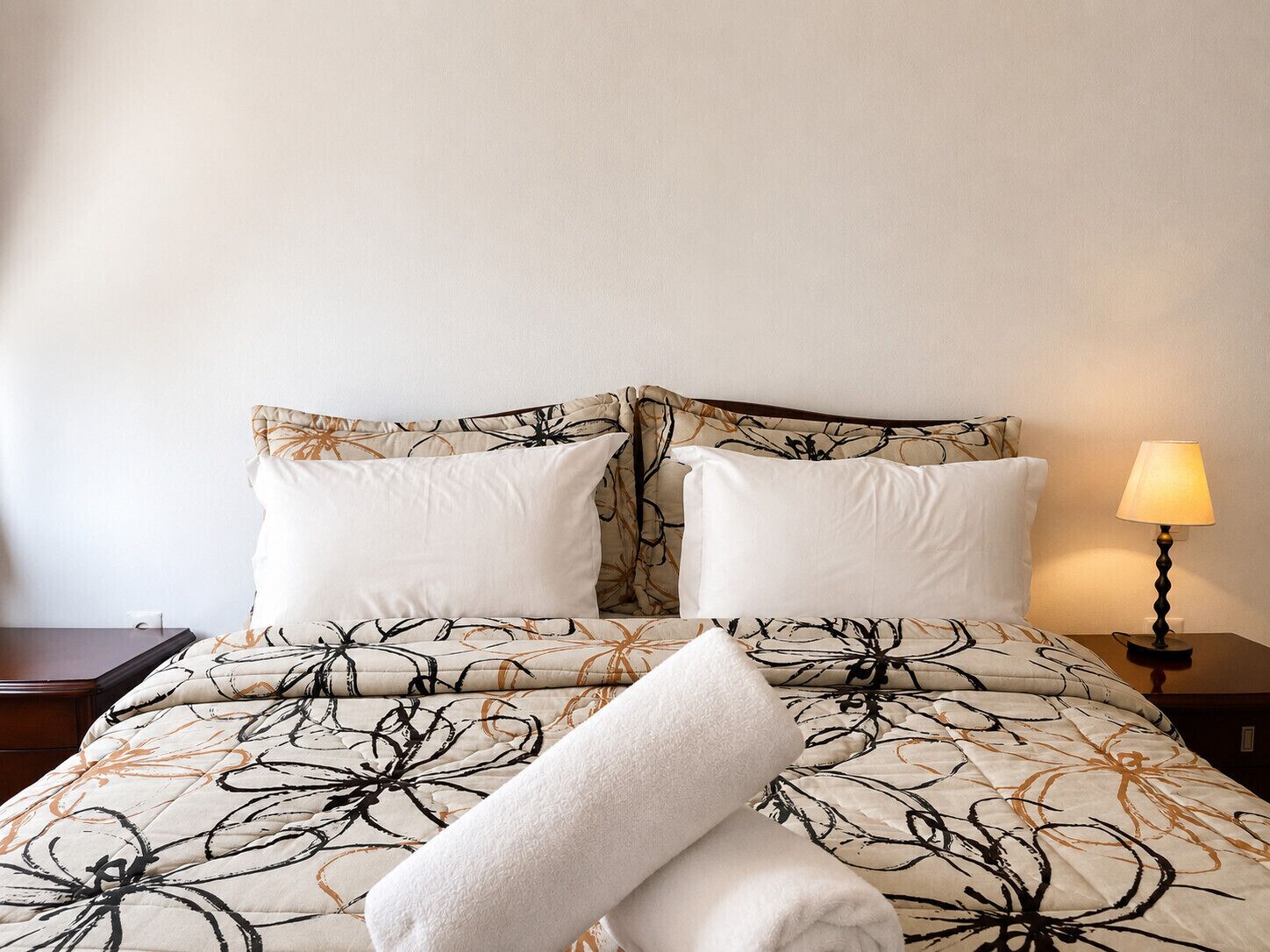 A neatly made bed with a floral-patterned duvet, two pillows, and a rolled white towel.