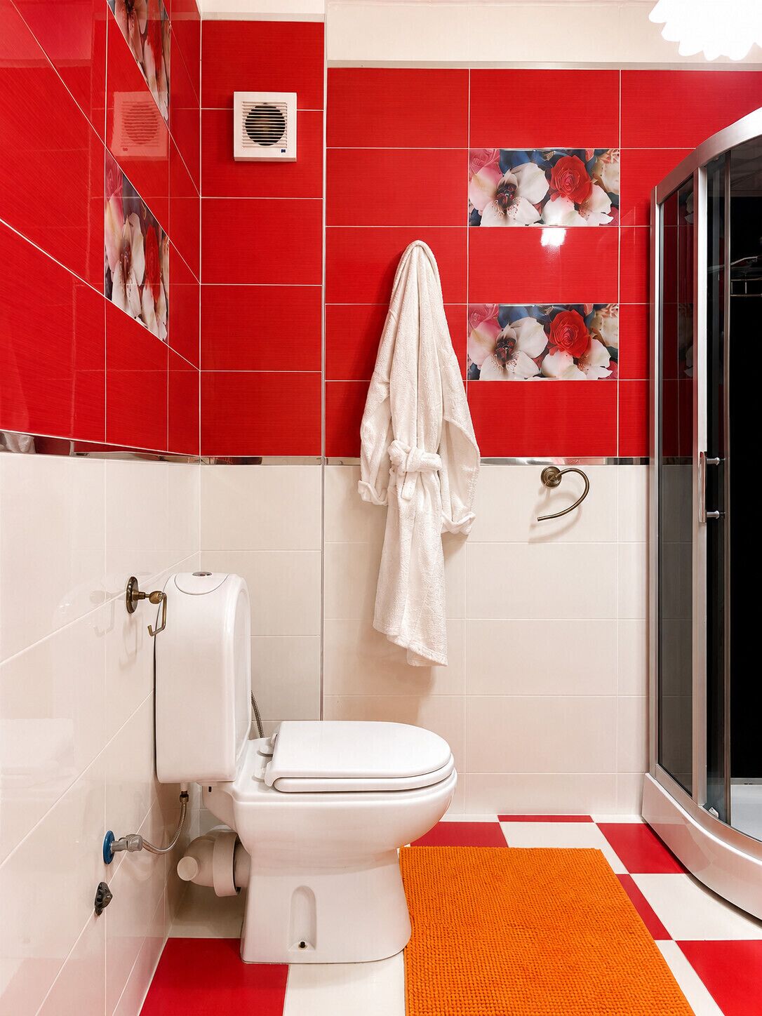 Et badeværelse med et hvidt toilet, en brusekabine i glas og en gulv med røde og hvide tern.