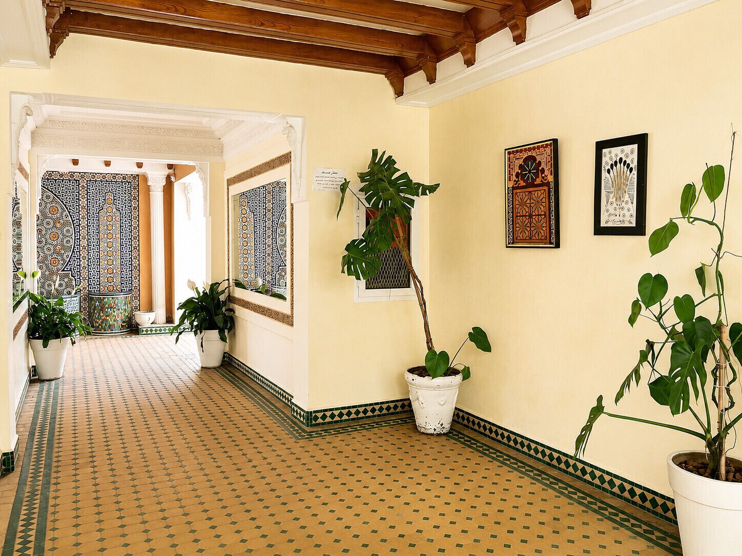 A hallway with patterned floor tiles, potted plants, and framed artwork on the walls.