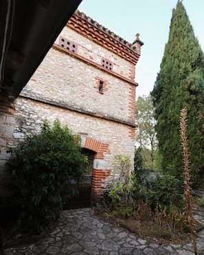 A stone building with a tiled roof, a narrow entrance, and a tall tree in the background.