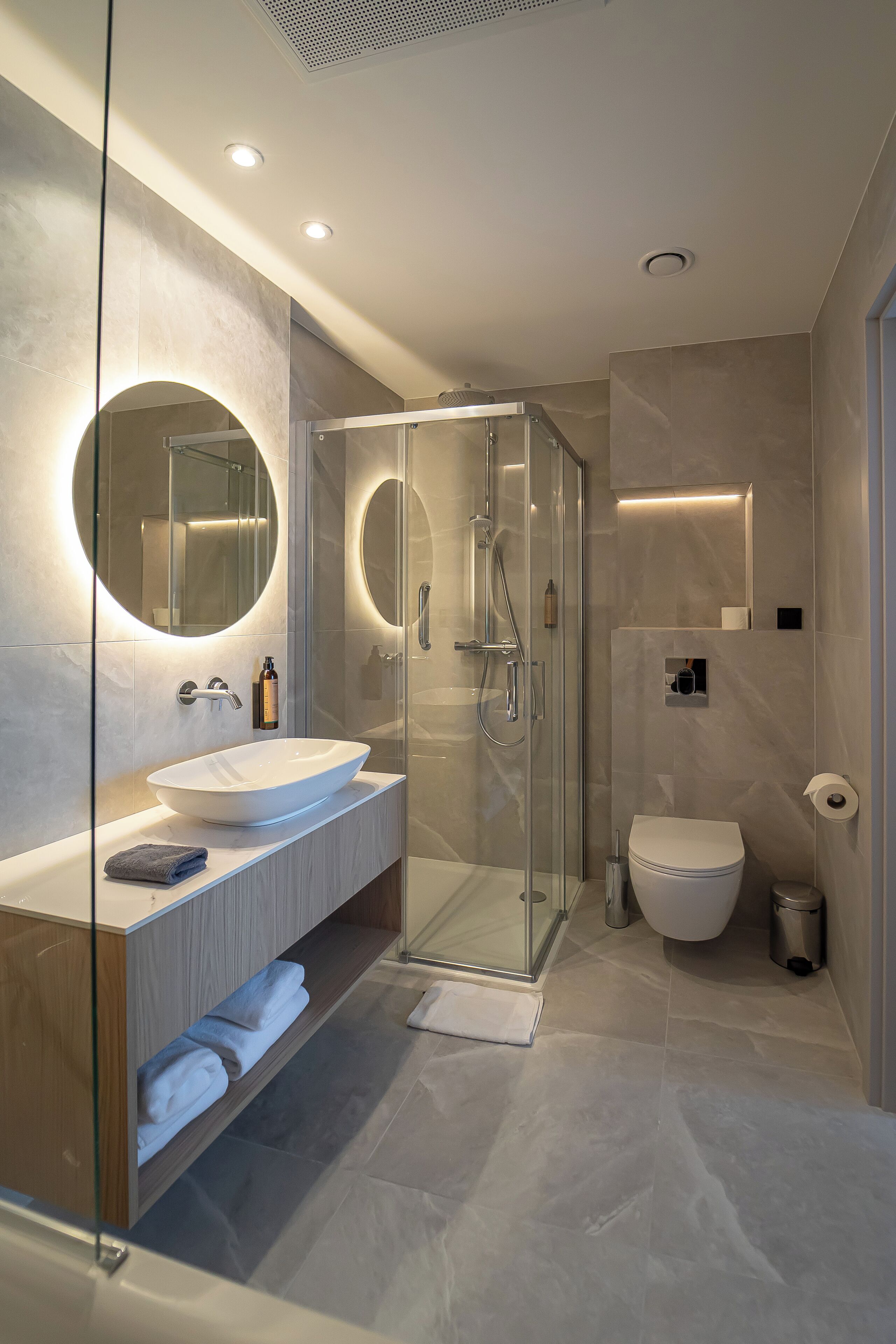 A modern bathroom with a glass-enclosed shower, a large round mirror, and a white sink.