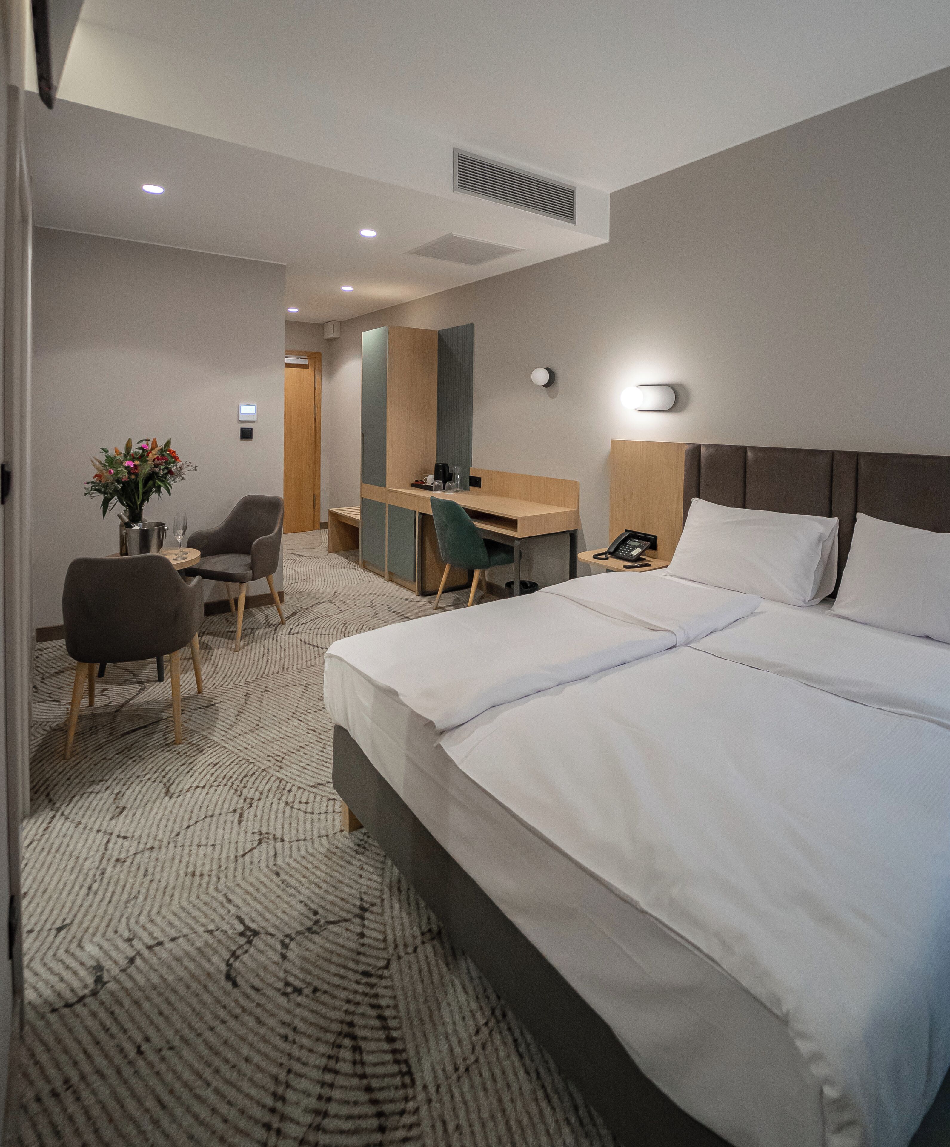 A hotel room with a large bed, a desk, a chair, and a small table with a vase of flowers.
