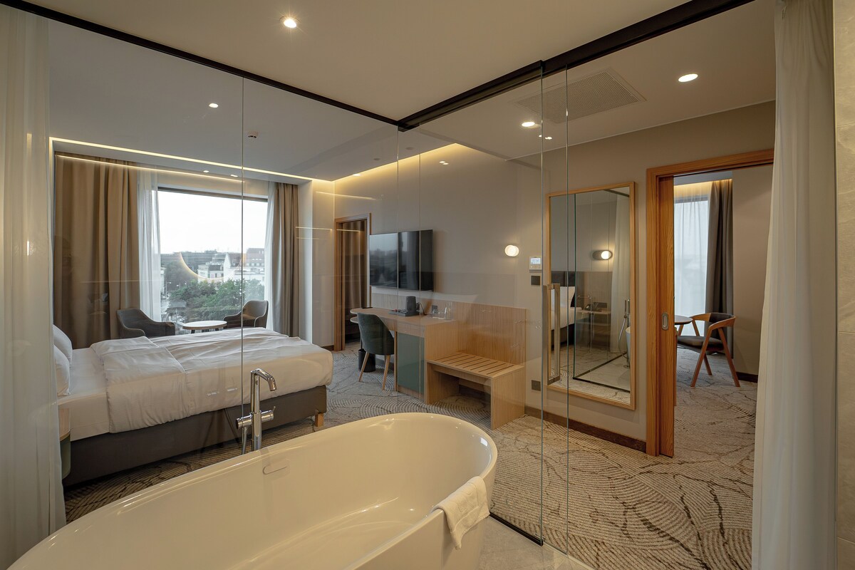 A modern hotel room with a large bed, a separate desk area, a glass-enclosed shower, and a view of the city.