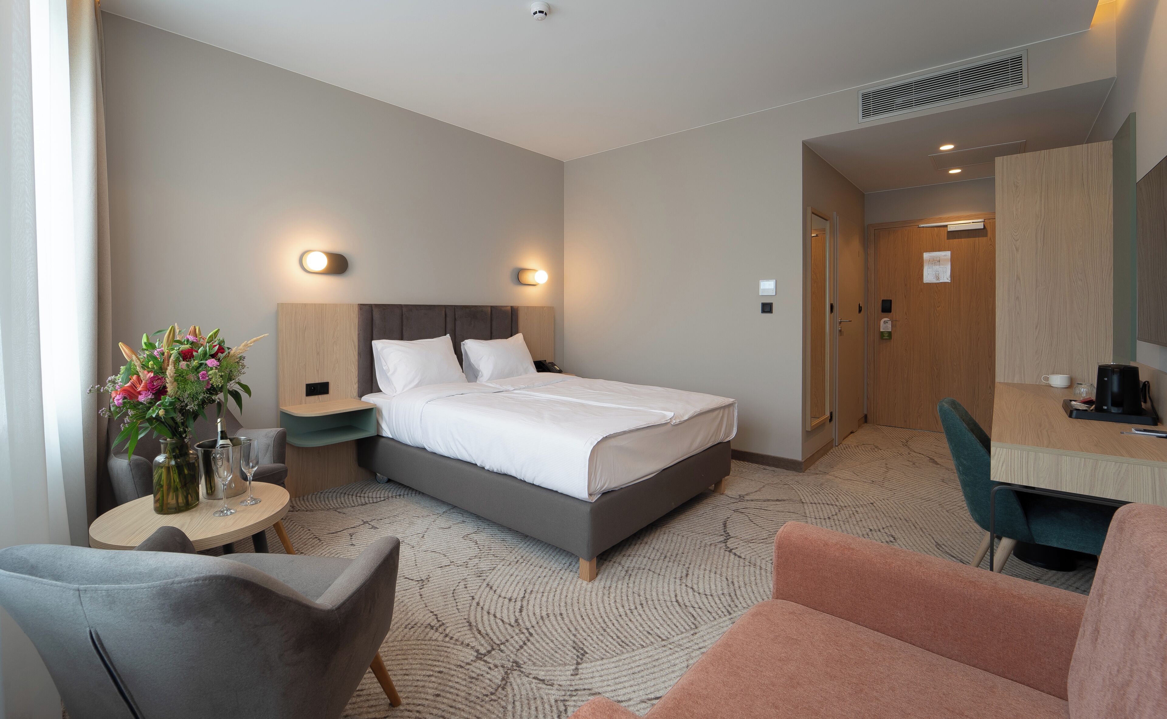 A hotel room with a bed, bedside tables, a desk, a chair, and a small table with a vase of flowers.